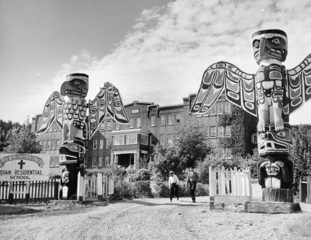 Black-and-white photograph of a Residential School. A ceremonial pole stands in the foreground.