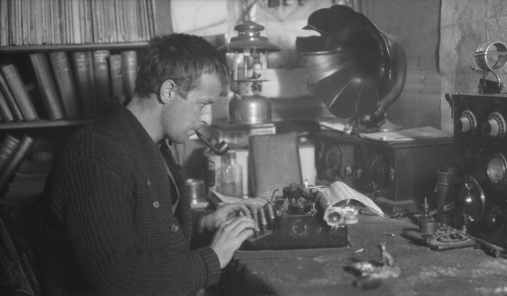 Photographie en noir et blanc d'un homme assis devant une machine à écrire, une pipe à la bouche.
