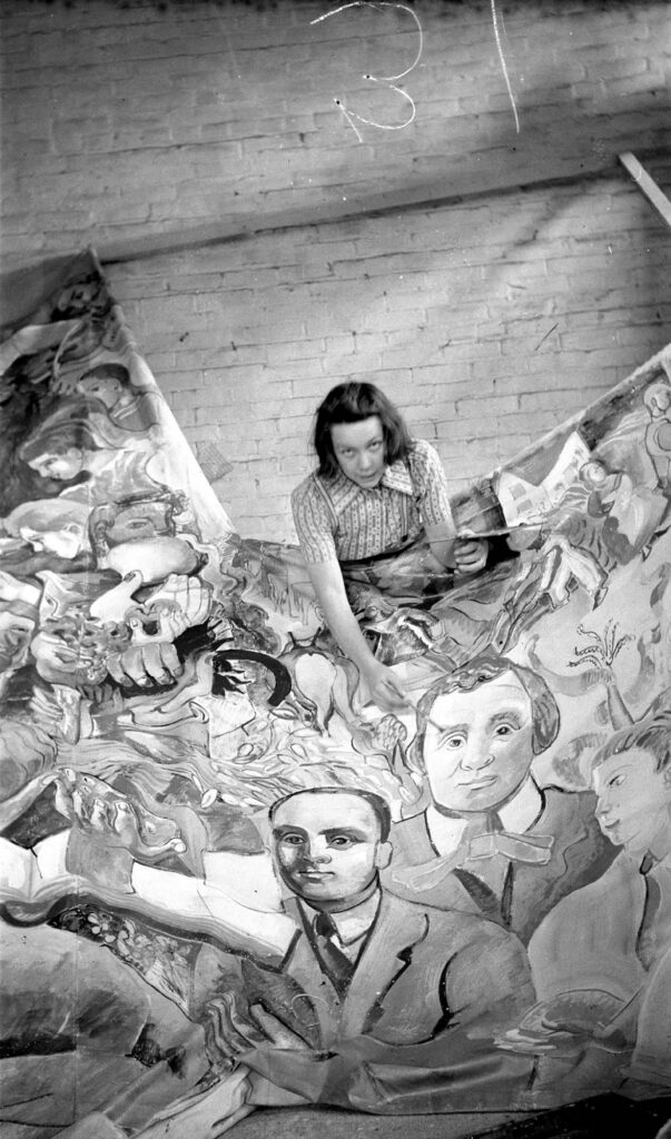 Photographie en noir et blanc d'une femme se tenant debout devant une grande fresque murale représentant de nombreux visages d'hommes, de femmes et d'enfants