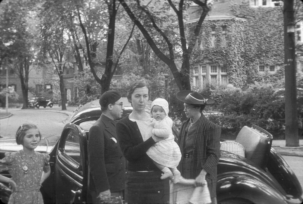 Photographie en noir et blanc représentant une jeune fille, un homme, une femme tenant un bébé dans ses bras et une autre femme, tous debout devant une voiture noire garée dans la rue.